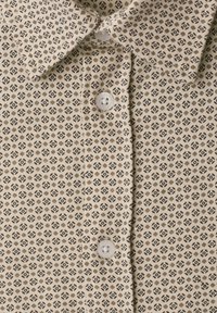 Chemise beige à boutons avec un petit motif géométrique répétitif en marron et noir et des boutons ronds blancs.