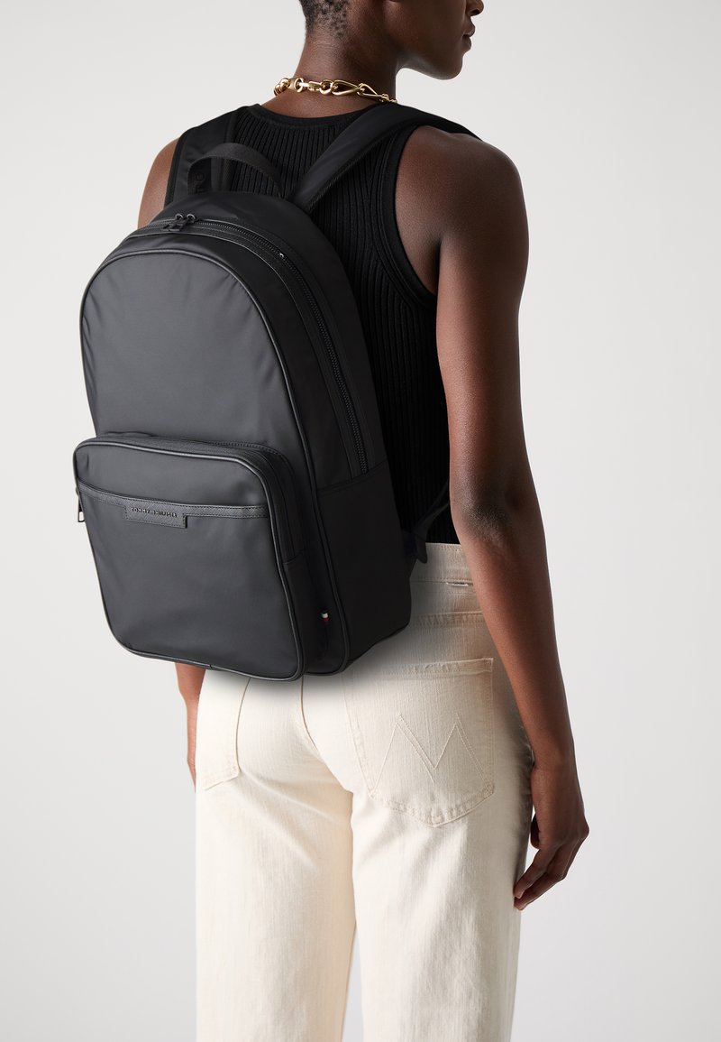 Black backpack with a smooth, matte finish. Features a front zipper pocket and padded shoulder straps. Worn on the back over light-colored pants.