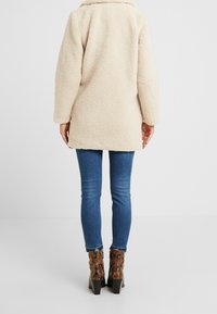 Persona con una chaqueta de forro polar beige, jeans ajustados azules y botas de tacón de tobillo marrones con un estampado de serpiente, de pie de espaldas.