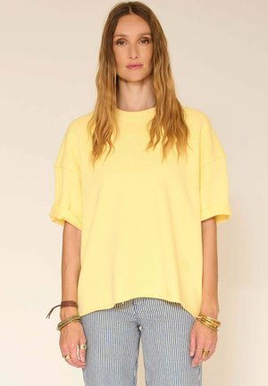 Femme aux longs cheveux ondulés portant un haut ample à manches courtes jaune et un pantalon rayé bleu et blanc sur un fond uni.