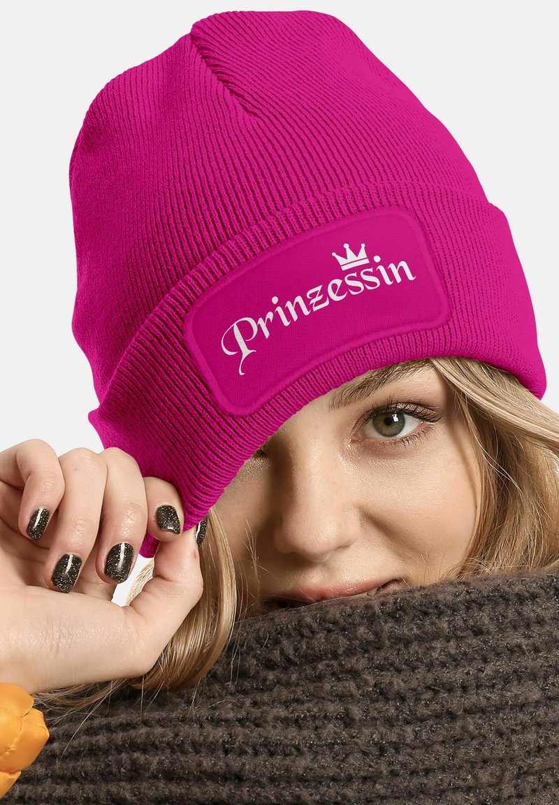 Bright pink knitted beanie with a textured finish. Features a white rectangular patch labeled "Prinzessin" with a small crown graphic.