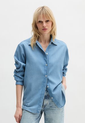 Femme blonde portant une chemise en jean bleu clair aux manches retroussées et un jean bleu délavé, debout avec une main dans la poche sur un fond blanc.