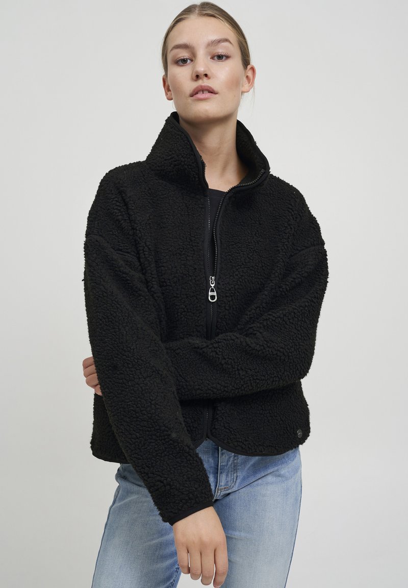 Black fleece jacket with high collar, zip closure, and textured fabric. Features long sleeves and a curved hemline. Paired with light blue jeans.
