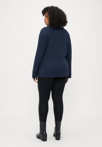 Haut à manches longues bleu marine avec une coupe droite, associé à un legging noir arborant un motif latéral, et des bottines noires à talons épais.