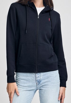 Osoba ubrana w czarną bluzę z kapturem na zamek, z przednimi kieszeniami i małym czerwonym logo na piersi, w połączeniu z jasnoniebieskimi jeansami.