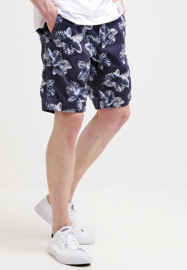 Homme portant un short bleu marine avec un imprimé floral blanc, baskets blanches, mains dans les poches, fond blanc.