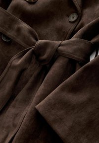 Cappotto in suede marrone con una cintura legata in vita, due bottoni e maniche ampie. Il tessuto presenta un aspetto morbido e testurizzato.