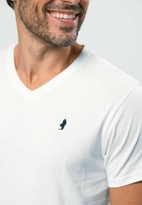 Baltas medvilninis V formos iškirptės marškinėlis su mažu tamsiai mėlynu išsiuvinėtu logotipu krūtinės srityje. Minkšta tekstūra ir laisvas siluetas.