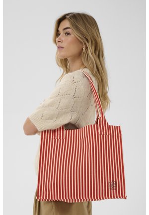 Mlada ženska z dolgimi blond lasmi, oblečena v kremno pleteno majico, nosi rdečo in belo navpično črtasto torbo čez ramo.