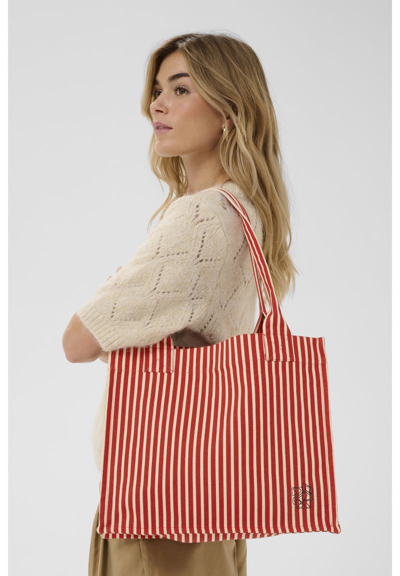 Jauna moteris su ilgais blondiniais plaukais, dėvinti kreminės spalvos megztinį, ant peties nešiojanti raudonai baltą vertikaliai dryžuotą krepšį.