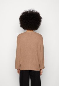 Person mit Afro-Frisur, die einen lockeren braunen Strickpullover und schwarze Hosen trägt und von einer schlichten weißen Wand abgewandt ist.