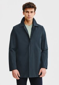 Granatowy wodoodporny płaszcz z kołnierzem, pełnym zapięciem na zamek z przodu oraz długimi rękawami. Gładka tekstura i dopasowany krój. Noszony na zielonej koszuli.