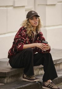 Kurtka z polaru w bordowo-kremowy wzór kwiatowy, czarne spodnie szerokie nogawki oraz brązowe trampki z czarnymi paskami, trzymająca filiżankę kawy.