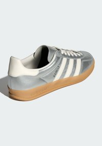 Silver Adidas Gazelle-sneakers med vita ränder, beige hälla, vita snören och sula i gummi på en ljus bakgrund.