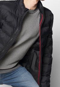 Black puffer jacket with a zip closure and red interior. Underneath, a patterned gray sweater featuring a ribbed neckline and hem.