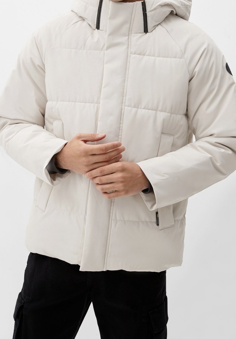 Homme portant une veste d'hiver matelassée beige clair, les mains jointes devant, pantalon cargo noir visible en bas.