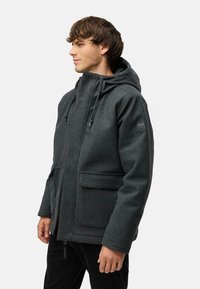 Mørkegrå hættejakke lavet af fleece, med lynlås lukning, to frontlommer, justerbare snore og et diskret logo patch på ærmet.