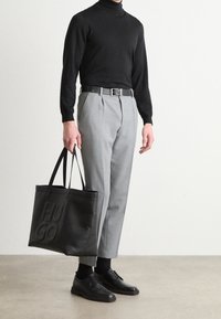 Black turtleneck sweater, gray pleated trousers, black leather belt, and black leather shoes. Holds a large black tote with "HUGO" embossed.