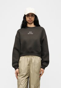 Γυναίκα με μακριά σκούρα μαλλιά που φοράει λευκό καπέλο LA, σκούρο γκρι φούτερ New Balance και μπεζ παντελόνι cargo, στέκεται μπροστά σε απλό φόντο.