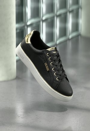 Schwarzer Guess Sneaker mit goldenen Akzenten und Logomuster, weiße Sohle, schwebend über poliertem Boden mit Glasblock-Hintergrund dargestellt.