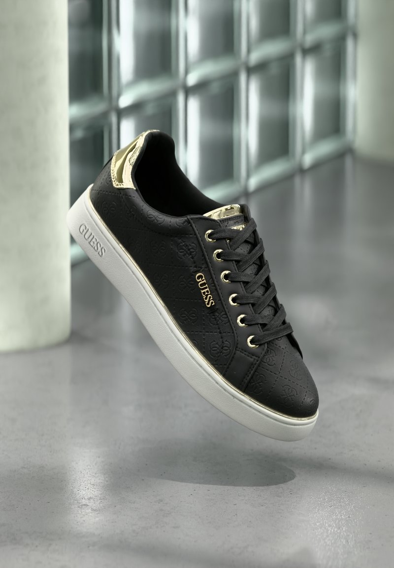 Baskets Guess noires avec accents dorés et motif logo, semelle blanche, présentées en suspension au-dessus d'un sol poli avec un fond de blocs de verre.