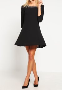 Robe noire à jupe évasée et manches trois-quarts. Présente un col orné de bijoux. Tissu lisse, longueur au-dessus du genou, associée à des escarpins noirs.