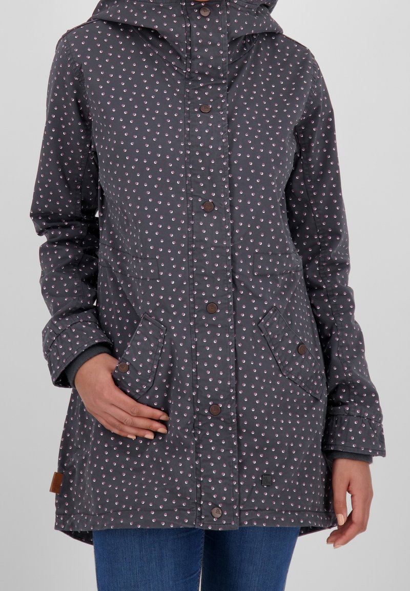 Imperméable à capuche gris foncé avec petits pois roses et blancs, fermetures à boutons, poches à rabat à l'avant, porté avec un jean bleu.