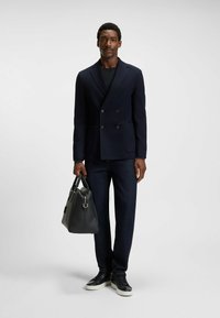 Navy double-breasted blazer with textured fabric, paired with dark jeans and a black leather handbag. White-soled black sneakers complete the outfit.