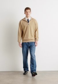 Pullover beige a maniche lunghe con colletto a metà zip indossato sopra una camicia bianca e una cravatta blu navy; abbinato a jeans blu e scarpe nere.