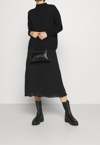 Schwarzer Strickpullover mit hohem Kragen, schwarzer plissierter Midirock, schwarze Stiefeletten und eine kleine schwarze Handtasche mit strukturierter Oberfläche.