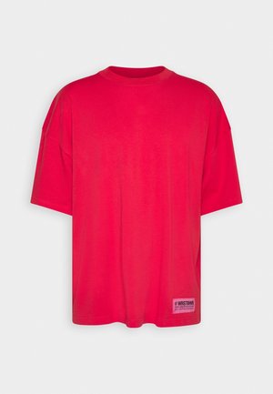 T-shirt rouge surdimensionné avec un col rond, des manches courtes et une étiquette texturée sur le bas devant. Confectionné en tissu de coton lisse.