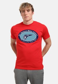 Rød kortærmet T-shirt lavet af bomuld med et blåt oval grafisk design, der viser "Pan American World Airways" og en globe.