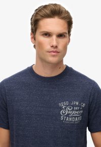 Joven con cabello castaño claro que lleva una camiseta azul marino con el texto blanco "Super Dry Standard" en el bolsillo, mirando de frente contra un fondo blanco.