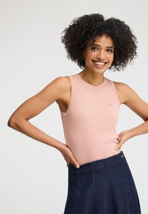 Femme souriante aux cheveux bouclés portant un haut côtelé rose sans manches et une jupe bleu foncé taille haute, les mains posées sur les hanches.