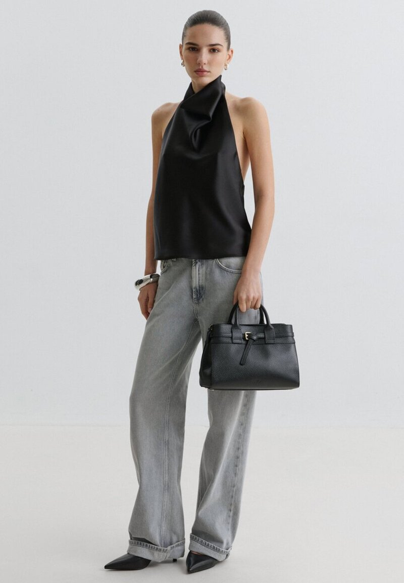 Femme en top noir à dos nu et jean gris large tenant un sac à main en cuir noir devant un fond clair uni.
