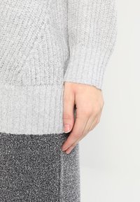 Ljust grå stickad tröja med ribbat nederkant och ärmar, med ett strukturerat mönster. Handen vilar mot ett mörkgrafitgrått, glittrigt tyg.