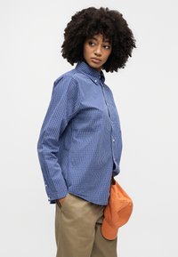 Γυναίκα με σγουρά μαλλιά που φορά μπλε ριγέ πουκάμισο με κουμπιά και μπεζ παντελόνι, κρατώντας ένα πορτοκαλί καπέλο στο χέρι της.