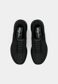 Scarpe sportive nere con tomaia testurizzata, design con lacci e sottopiede imbottito, etichetta con scritto "Skechers Slip-Ins" all'interno.