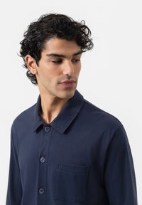 Chemise bleu marine à manches longues avec col, dotée d'une seule poche poitrine et de boutons violets. Tissu lisse avec une texture subtile.