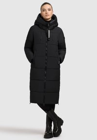 Sort pufferjakke med hætte, der har lynlås, forlommer og et langt, quiltet design. Kombineret med kraftige sorte støvler.