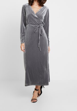 Femme portant une robe portefeuille en velours gris argenté à manches longues avec une ceinture à la taille, associée à des sandales à talons hauts noires avec bride à la cheville.