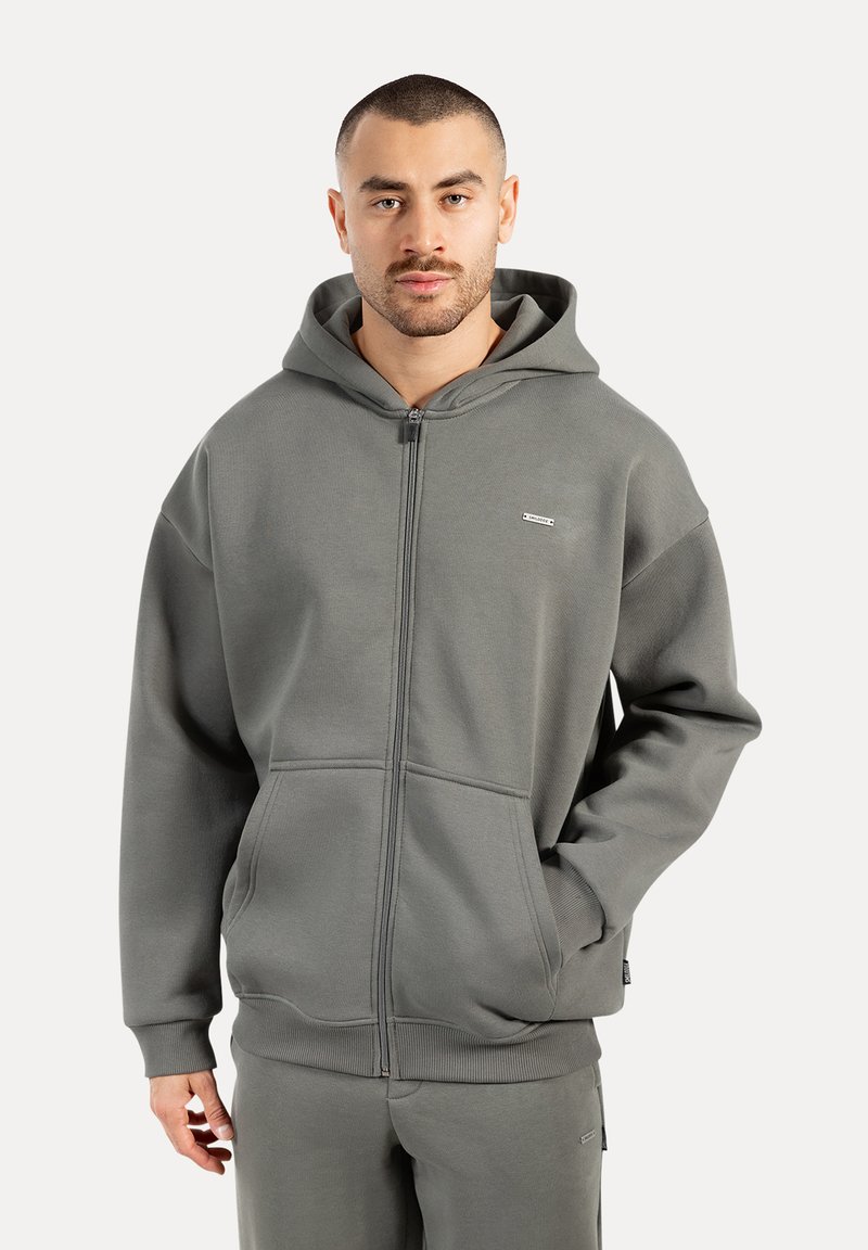Homme portant un sweat à capuche gris avec zip, poches avant et un pantalon gris assorti devant un fond uni.