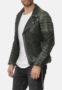 Giacca da motociclista in pelle verde con design aderente, spalle trapuntate, cerniere argentate e zip laterali. Indossata sopra una t-shirt bianca.