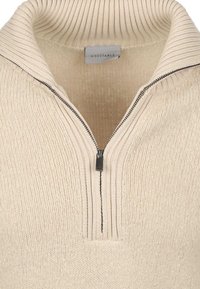 Beige gebreide trui met een halve rits en een geribbelde omgeslagen kraag, met zichtbare textuur en merkembleem aan de binnenkant van de kraag.