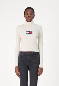 Maglione a collo alto a coste crema con un patch ricamato in blu, rosso e bianco sul petto, abbinato a jeans in denim scuro.