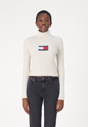 Maglione a collo alto a coste crema con un patch ricamato in blu, rosso e bianco sul petto, abbinato a jeans in denim scuro.