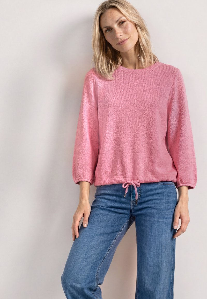 Femme aux cheveux blonds portant un pull rose à manches longues avec un cordon à l'ourlet et un jean bleu, debout contre un mur clair uni.