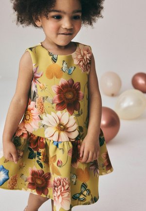Junges Kind mit lockigem Haar, das ein gelbes Kleid mit Blumenmuster trägt und neben roségoldenen und cremefarbenen Luftballons auf hellem Hintergrund steht.
