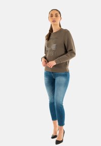 Felpa verde oliva scuro con testo grafico metallico, abbinata a jeans skinny blu chiaro e tacchi alti neri.