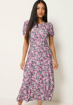 Kvinde står iført en pink, lilla og grøn blomster-middelængde kjole med korte puffærmer og ser fremad mod en ensfarvet baggrund.